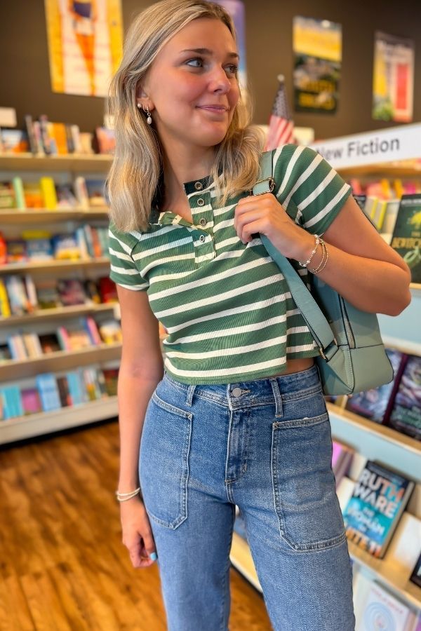 Easy A Green Striped Baby Tee with cropped fit and V-neck button detail, styled with high-waisted jeans in a bookstore setting.