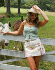 Sage Green Halter Snap Front Crop Tank styled with a printed mini skirt and cowboy hat, perfect for casual outdoor fashion.
