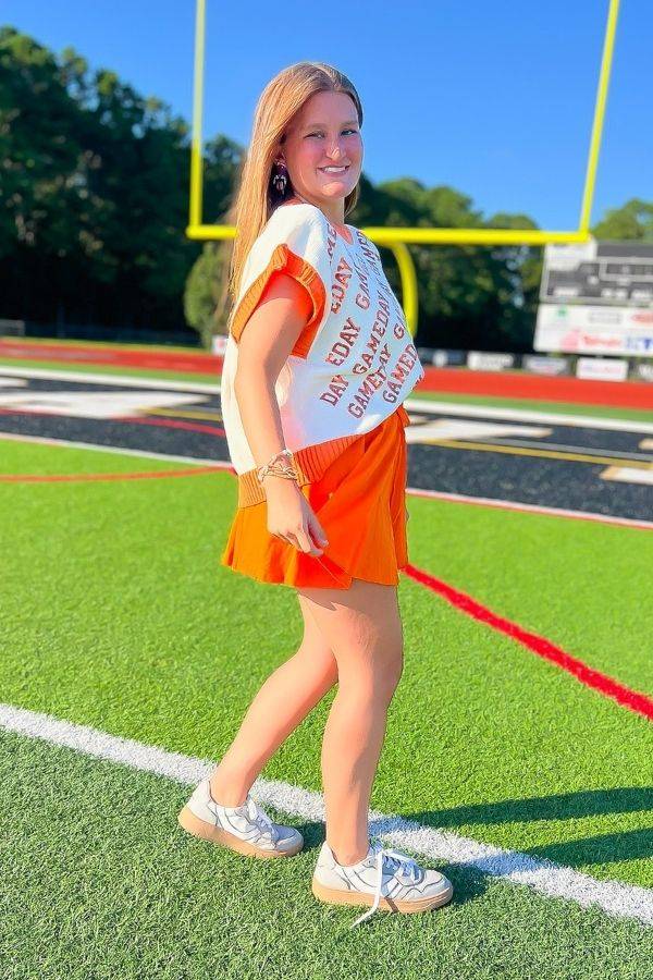 Model walking on football field in GAMEDAY Sequin Sweater Vest - Orange & White. Sleeveless knit vest styled for gameday fashion.