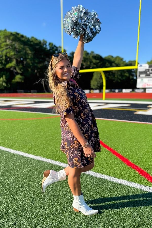 Cheerful model in Tiger Printed Button Down Mini Dress holding pom-poms, styled for gameday with white boots and a sporty vibe.