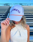 Clemson White Mesh Back Trucker Hat with upside-down purple embroidery and orange rope detail, styled for game day in stadium bleachers.