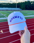 Clemson White Mesh Back Trucker Hat – Snapback with orange rope and bold purple lettering, held in front of a football field for gameday flair.
