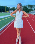 Model poses in Ellie Beige Halter Neck Scalloped Mini Dress with halter tie and boots, styled for a sporty game day look on the track.
