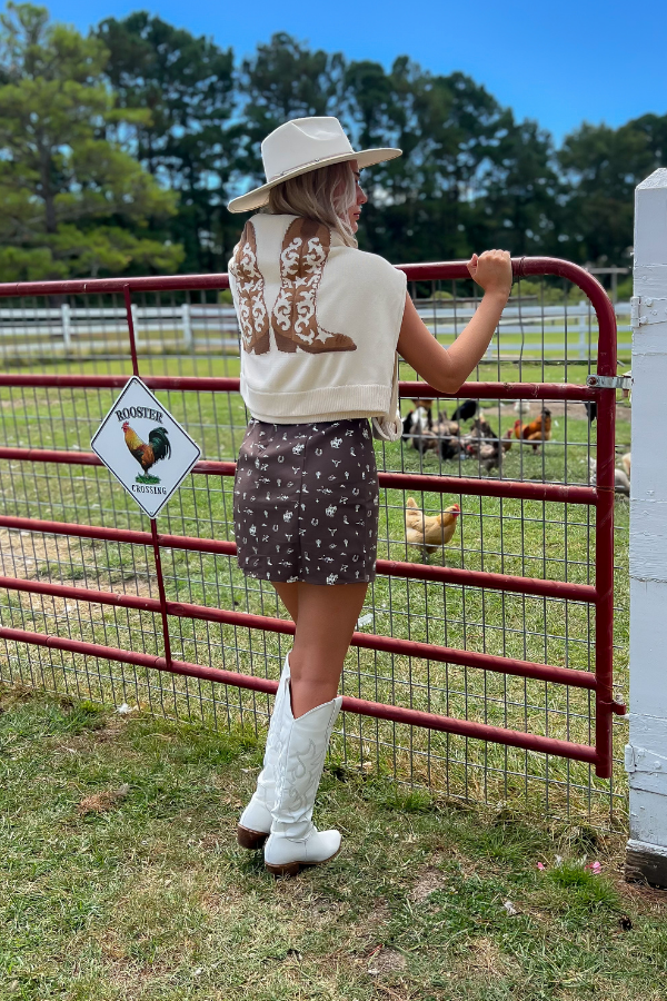 Back view of Show Me Your Mumu Cowboy Boot Knit Stay Awhile Sweater draped over shoulders, paired with a brown mini dress and white cowboy boots for a western-inspired outfit.
