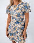 Close-up of floral dress with puff sleeves, beige background, and blue flower pattern, styled with jewelry