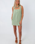 Side view of the green gingham mini dress with sleeveless design, pockets, and front buttons, paired with tan platform sandals