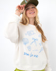 Sea La Vie Beach Icons Sweatshirt styled with green satin shorts and a green cap, showcasing relaxed beachwear fashion.