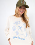 Model wearing Sea La Vie Beach Icons Graphic Sweatshirt with casual green shorts and a trucker hat, perfect for laid-back beach style.
