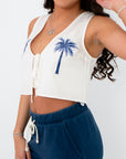 Woman wearing a white sleeveless top with palm tree design and blue pants on a white background