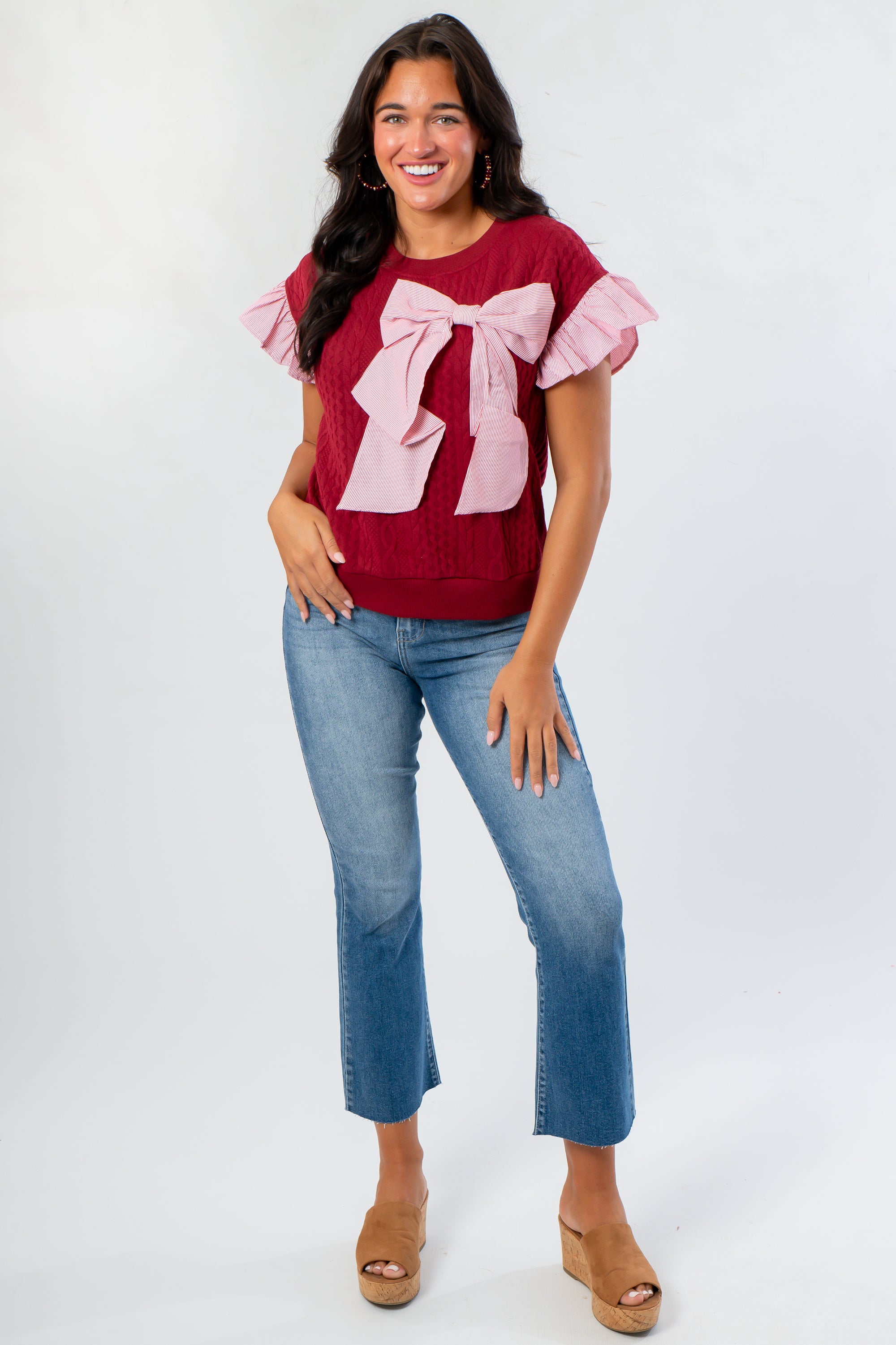 Burgundy Bow Cable Knit Top with pink ruffle sleeves and oversized bow, styled with cropped jeans and wedge sandals for a cozy chic look.