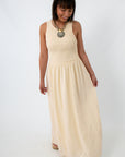 Side view of woman in cream maxi dress with smocked top and flowing skirt, smiling and looking down