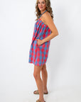 Side view of woman in a red and blue plaid mini dress with adjustable straps, smiling and looking down, white background