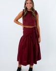 A woman modeling a matching burgundy two-piece set consisting of a sleeveless crop top with shoulder ties and a high-waisted wrap midi skirt, paired with black platform sandals, against a plain white studio background.