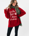 Show Me Your Mumu Twas The Night Knit Classic Crewneck Sweater in red with festive holiday graphic, styled with black leggings and plaid scarf.