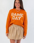 Game Day Orange Knit Sweater with bold white block letters, styled with a beige skirt for a cozy fall game day look.