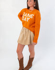 Side view of Game Day Orange Knit Sweater styled with a pleated beige skirt and western boots, ideal for fall gameday outfits.