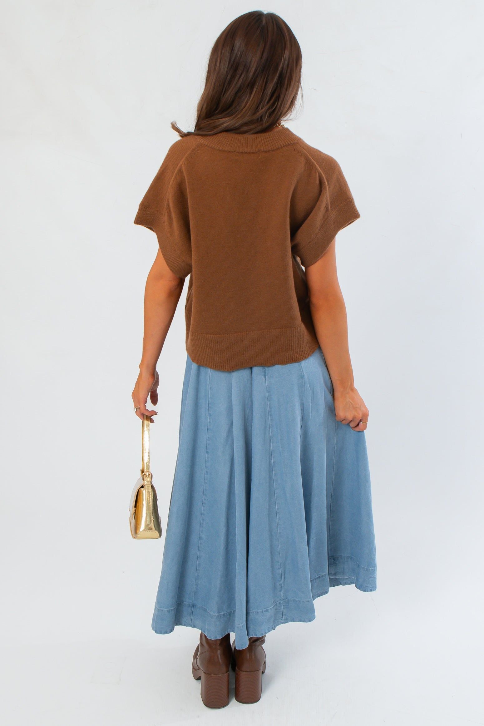 Back view of light brown V Neck Sweater Vest with ribbed hem, worn with a flowy blue skirt and brown platform boots.