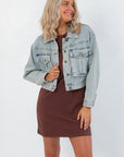 Maci Medium Wash Crop Denim Jacket – Classic & Trendy outerwear with front button closure and flap pockets, styled over a brown dress.