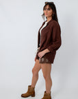 Women’s brown button-up shirt with long sleeves, layered over a white top, paired with embroidered shorts and cowboy boots, side view