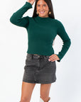 Taylor Emerald Green Ribbed Long Sleeve Top with mock neck and fitted silhouette, styled with a black denim mini skirt and white boots.