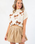Close-up of Football Bow Mock Neck Cream Sweater featuring brown football bow details and ribbed hem, styled with a tan mini skirt.