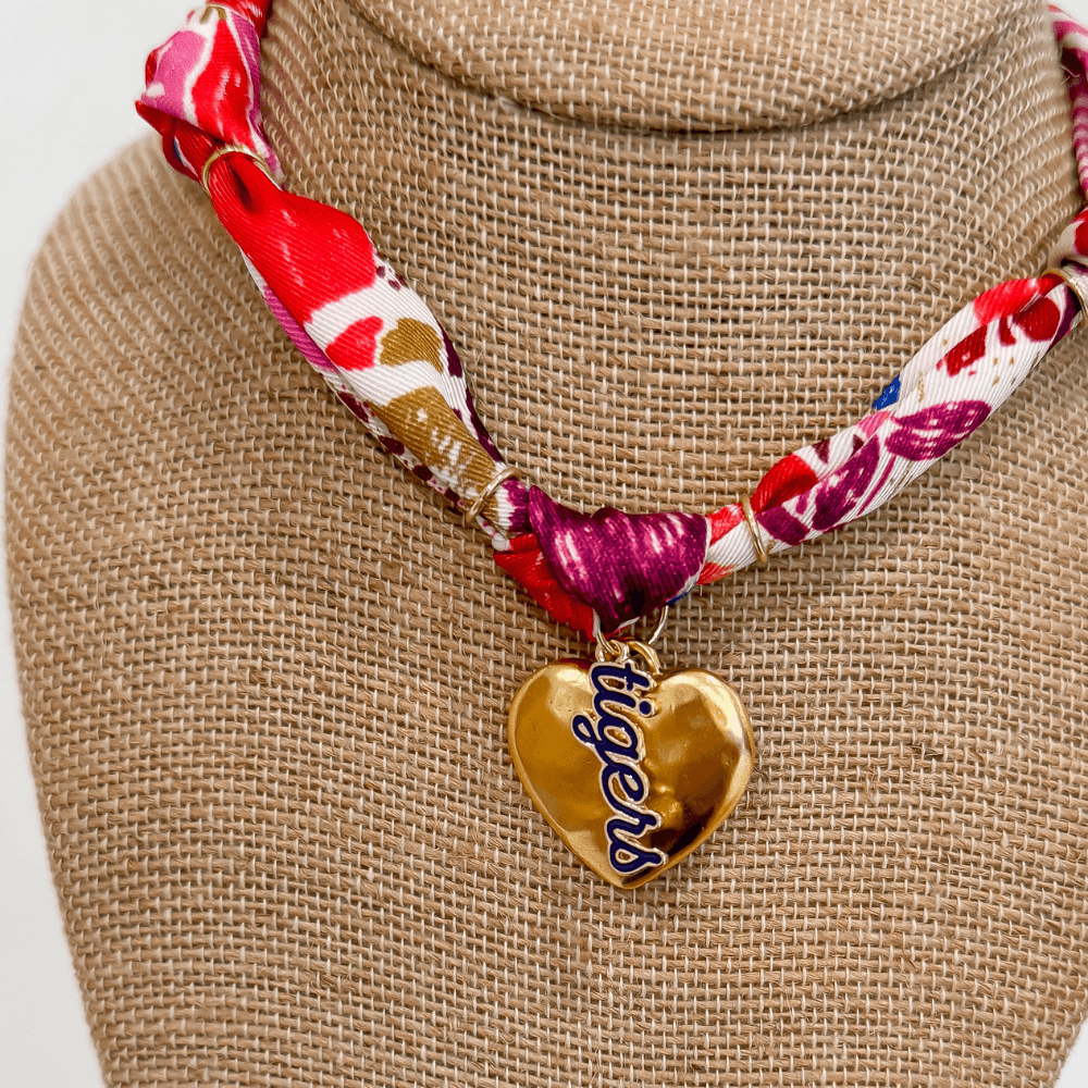 upclose photo of statement scarf necklace featuring gold heart pendant with Clemson tiger details and orange and purple abstract patterned knotted ribbon