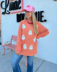 Orange Ribbed Sequin Ghost Sweatshirt – Festive Halloween Pullover with white sequin ghost graphics, styled with jeans and a pink cap.