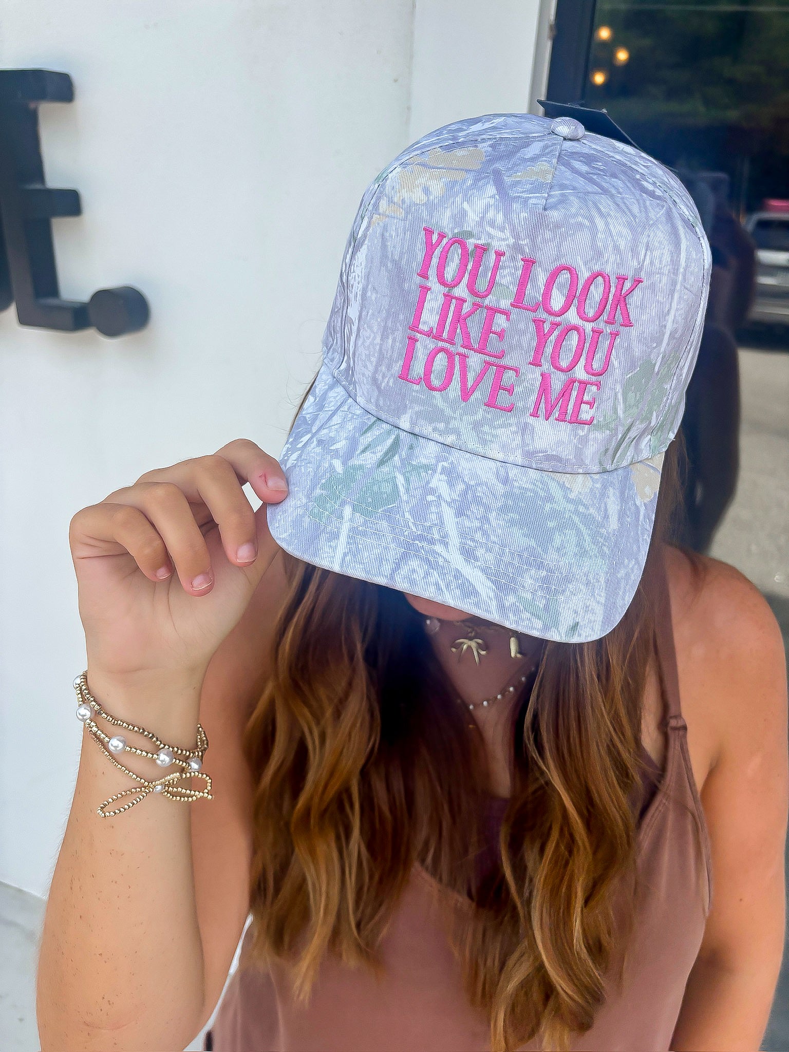 You Look Like You Love Me Camo Trucker Hat – Pink Lyrics Snapback worn by woman, featuring pink embroidered text on a gray camo background.