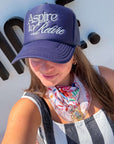 Woman wearing Aspire to Retire Navy Trucker Hat – Foam & Mesh Snapback with white cursive embroidery, styled with striped outfit and floral scarf.