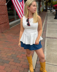 Woman in white top and blue shorts with yellow boots standing on a sidewalk with an American flag in the background.