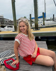 Woman in a red and white striped shirt and red shorts sitting on a dock with boats in the background.
