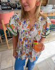 Front view of Spring Fling Floral Ruffle Sleeve Blouse featuring pleated details and vibrant floral pattern, styled with gold jewelry and a cocktail.