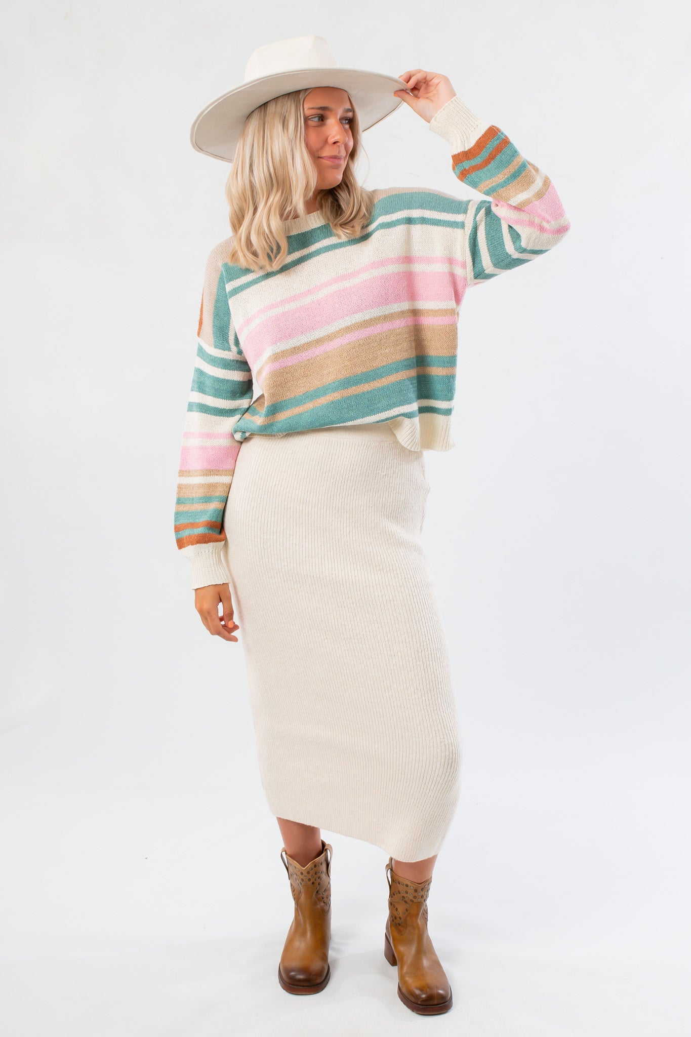 Woman wearing a colorful striped sweater, cream skirt, and cowboy boots on a white background