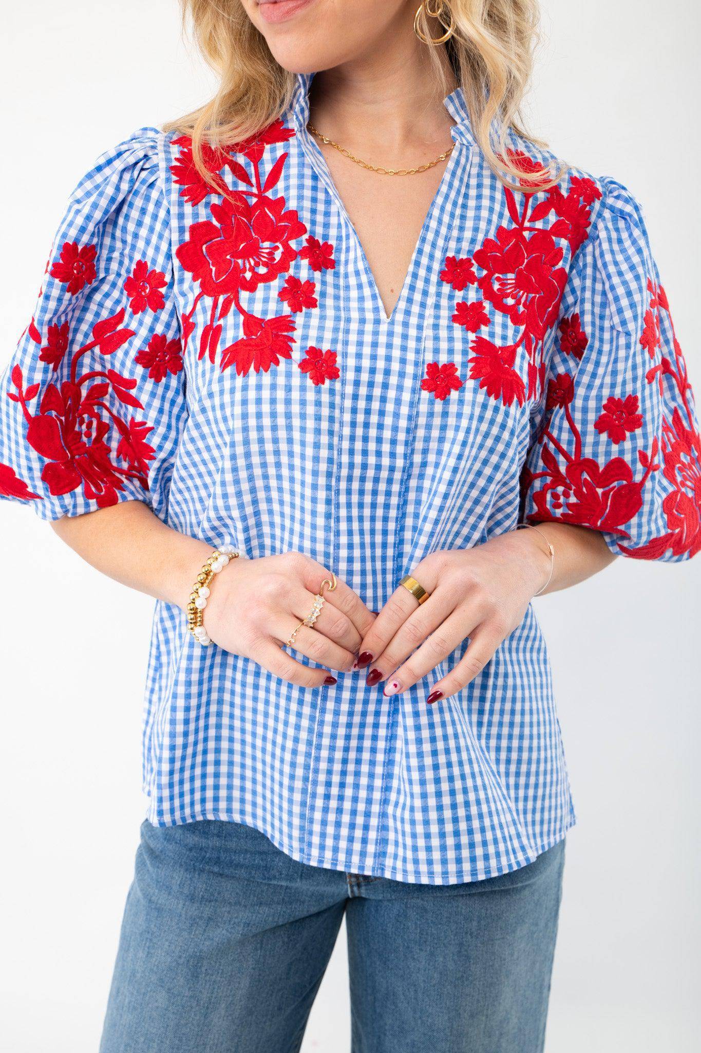 Close-up of THML Blue Gingham Embroidered Top LM showcasing intricate red floral embroidery on puff sleeves and a V-neckline, paired with gold accessories.