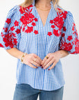 Close-up of THML Blue Gingham Embroidered Top LM showcasing intricate red floral embroidery on puff sleeves and a V-neckline, paired with gold accessories.