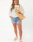 Model wearing Show Me Your Mumu Ciao Cocktail Woodsy Sweater with rolled denim shorts and accessories, showcasing cozy summer fashion.