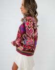 Side view of Chocolate Hearts Patterned Sweater featuring vibrant heart and geometric patterns on a brown base, paired with a white mini skirt.