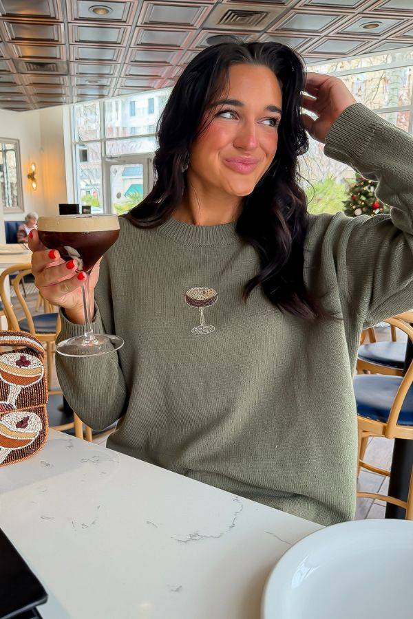 Woman wearing the Martini A Day Olive Beaded Sweater with espresso martini design, styled casually in a cafe setting. Cozy knitwear for holiday outings.