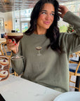 Woman wearing the Martini A Day Olive Beaded Sweater with espresso martini design, styled casually in a cafe setting. Cozy knitwear for holiday outings.