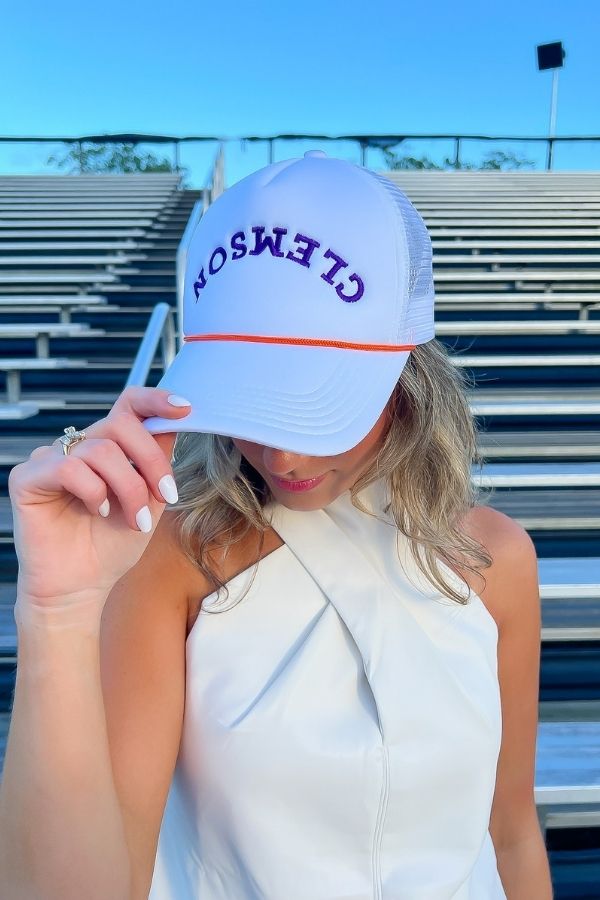 Clemson White Mesh Back Trucker Hat with upside-down purple embroidery and orange rope detail, styled for game day in stadium bleachers.