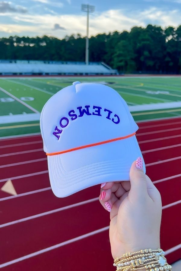 Clemson White Mesh Back Trucker Hat – Snapback with orange rope and bold purple lettering, held in front of a football field for gameday flair.