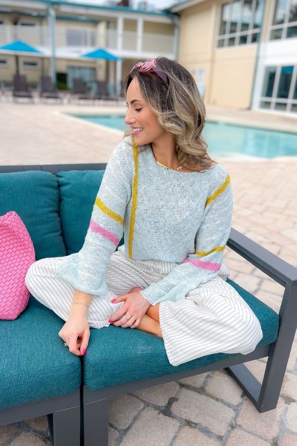 Baha Mint Stripe Beach Sweater with pastel pink, yellow, and ivory stripes, worn by a woman lounging by a pool. Lightweight and stylish for vacation wear.