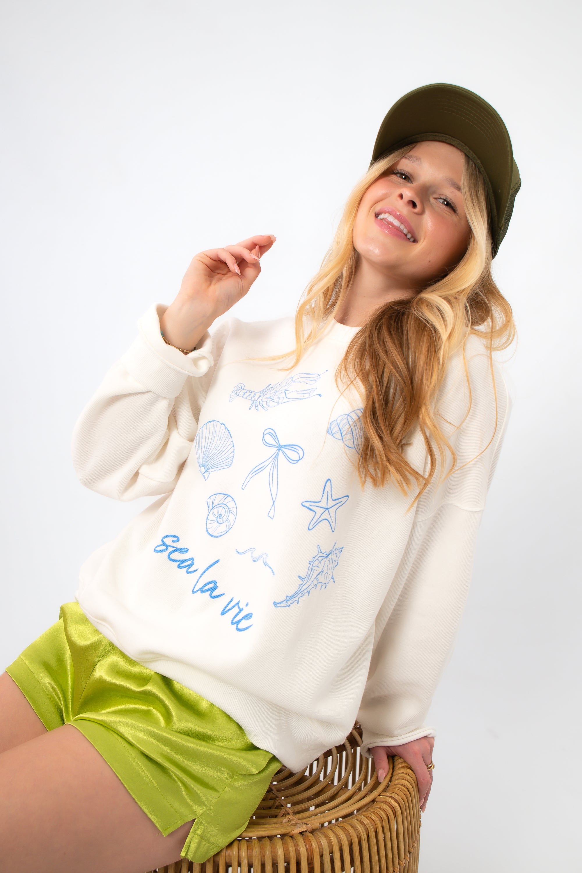 Sea La Vie Beach Icons Graphic Sweatshirt in white with blue seashell and starfish illustrations, styled casually with green shorts.