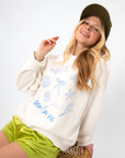 Sea La Vie Beach Icons Graphic Sweatshirt in white with blue seashell and starfish illustrations, styled casually with green shorts.