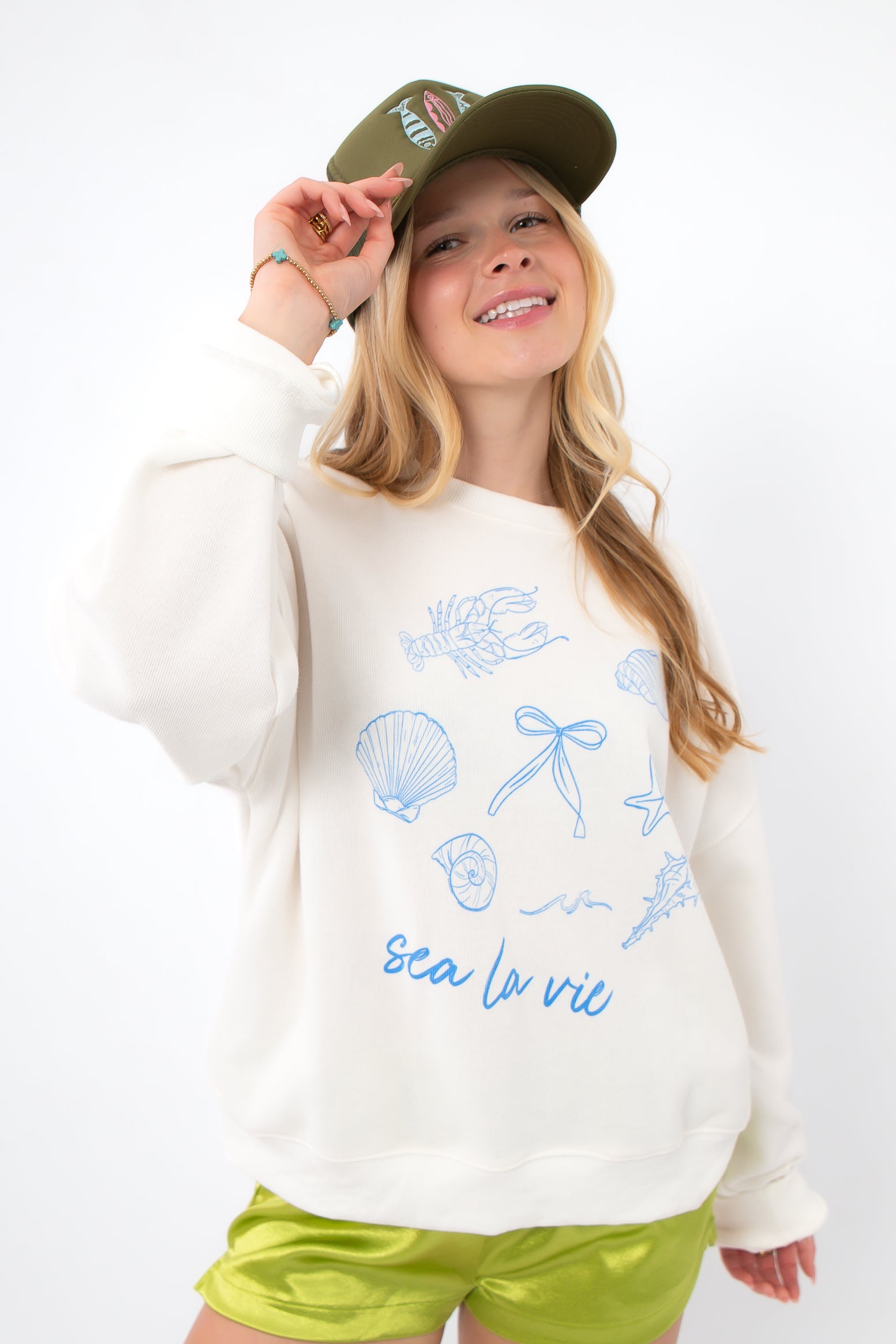 Sea La Vie Beach Icons Sweatshirt styled with green satin shorts and a green cap, showcasing relaxed beachwear fashion.