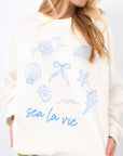Close-up of Sea La Vie Beach Icons Sweatshirt featuring detailed blue beach-themed graphics and 'sea la vie' text on soft white fabric.