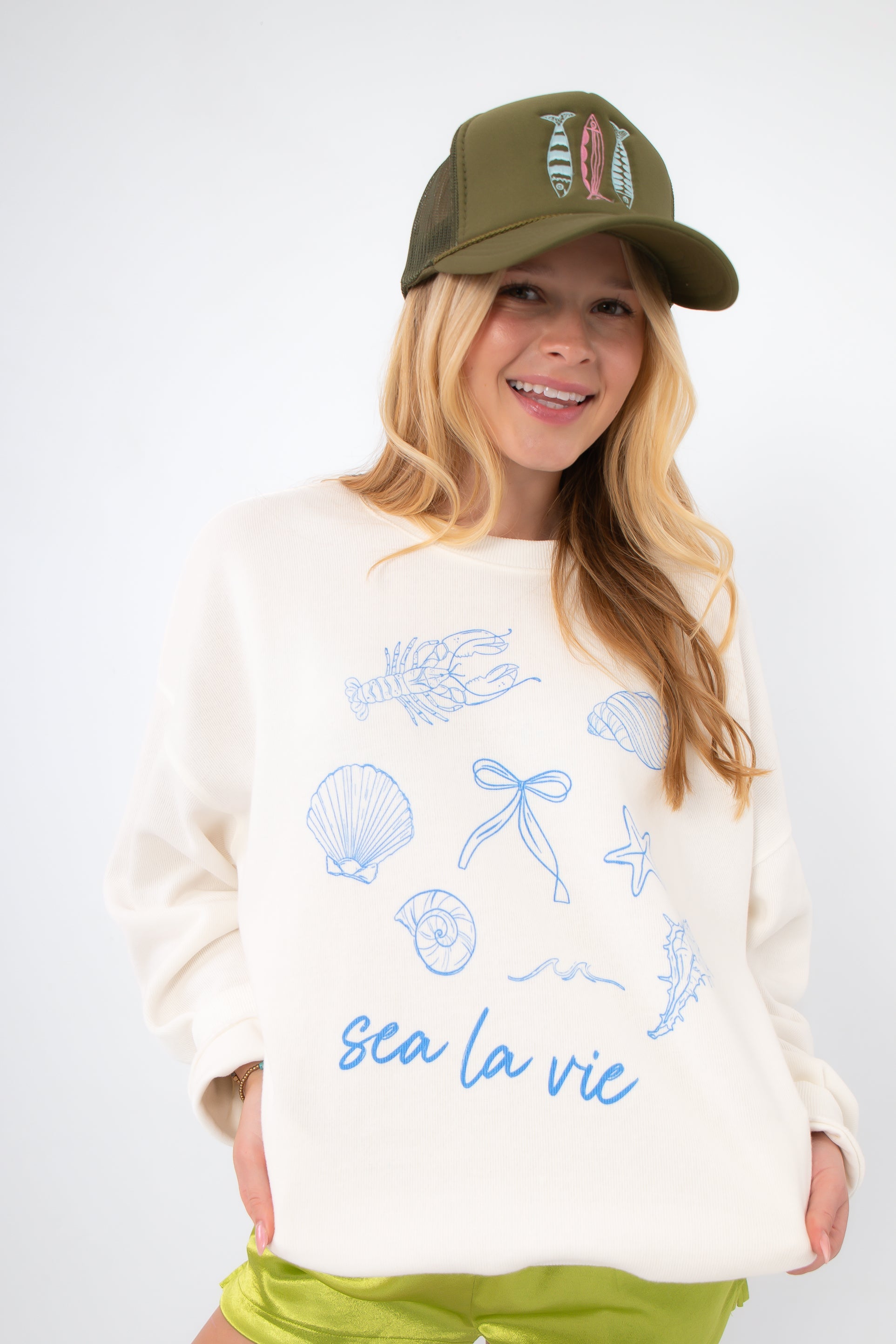 Model wearing Sea La Vie Beach Icons Graphic Sweatshirt with casual green shorts and a trucker hat, perfect for laid-back beach style.