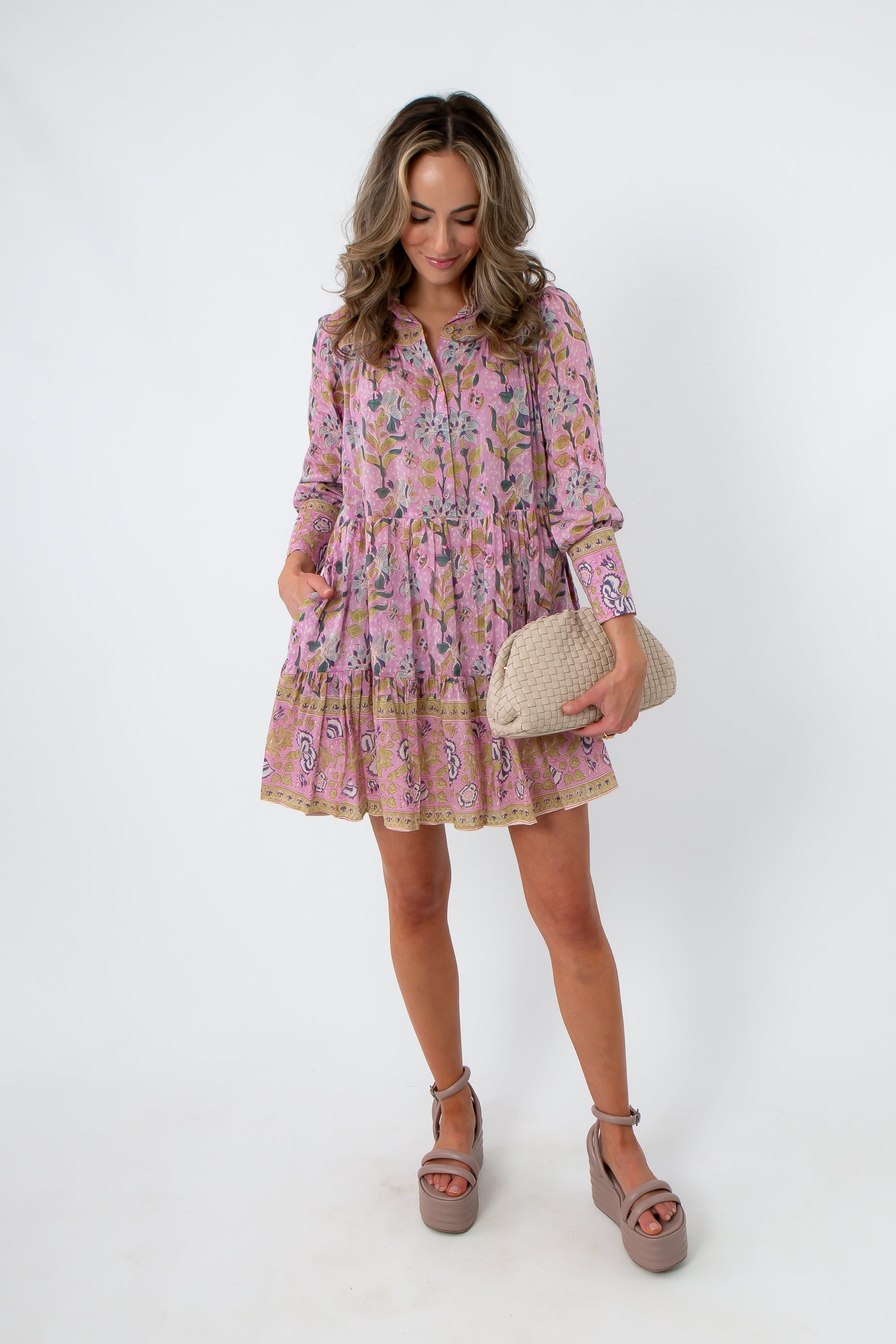 Pink and purple floral dress with tiered skirt, long sleeves, and pockets, styled with beige woven clutch and platform heels