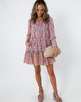 Pink and purple floral dress with tiered skirt, long sleeves, and pockets, styled with beige woven clutch and platform heels