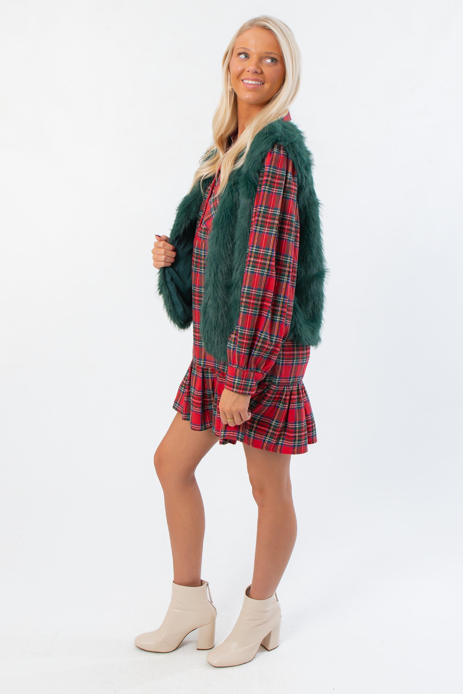 Side view of Frosted Pine Green Faux Fur Vest layered over a red plaid dress, showcasing cozy winter outerwear for women.